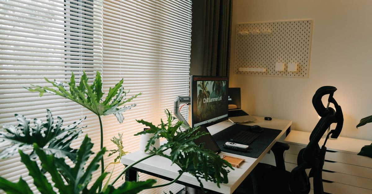 A stylish home office with a desk, chair, and lush green plants near a window with blinds.
