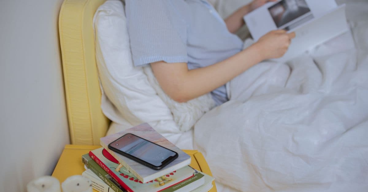 A woman enjoying a quiet moment reading in bed with books and a smartphone. Cozy indoor setting.